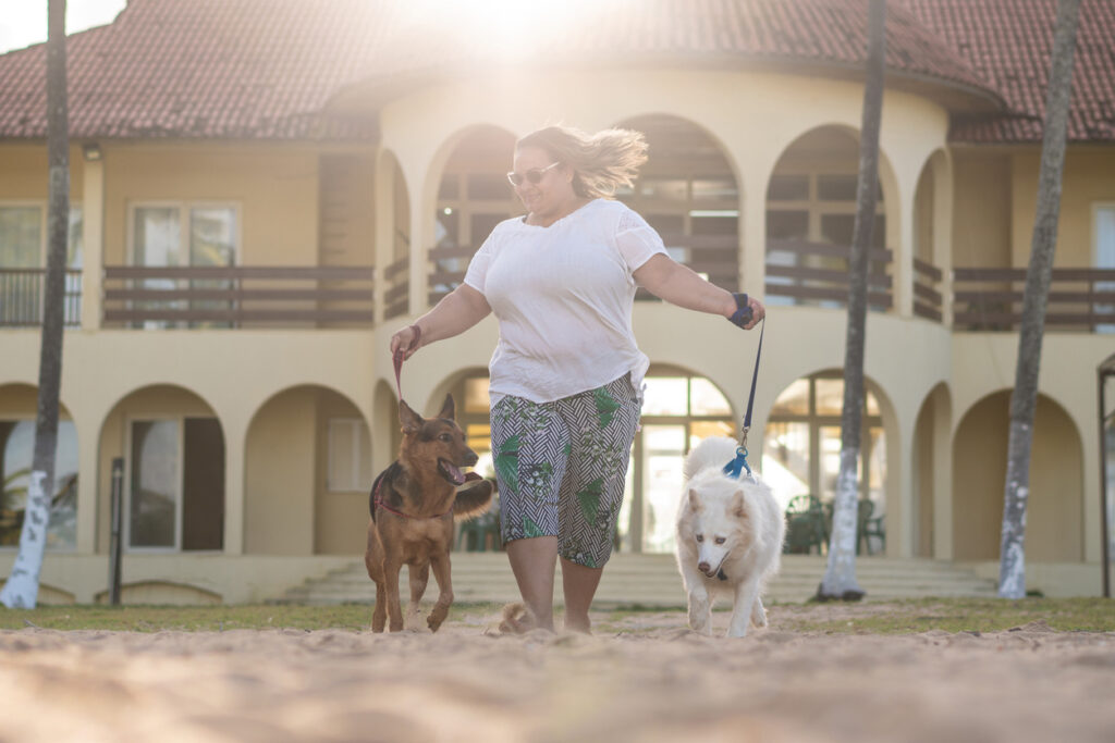A woman walks her two dogs on a beach in front of a large building with the sun behind them. 